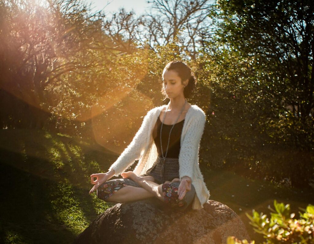 Woman in tranquil meditation pose outdoors, bathed in sunlight, surrounded by lush greenery.